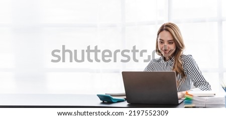 Young confident Asian businesswoman working at office desk and typing with a laptop, office shelves on background.