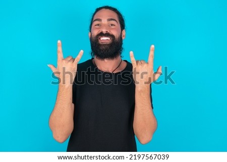 young bearded man wearing black T-shirt over blue studio background makes rock n roll sign looks self confident and cheerful enjoys cool music at party. Body language concept.
