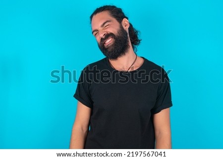 Positive young bearded man wearing black T-shirt over blue studio background with overjoyed expression closes eyes and laughs shows white perfect teeth