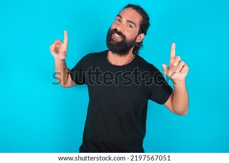 Photo of upbeat young bearded man wearing black T-shirt over blue studio background has fun and dances carefree wear being in perfect mood makes movements. Spends free time on disco party