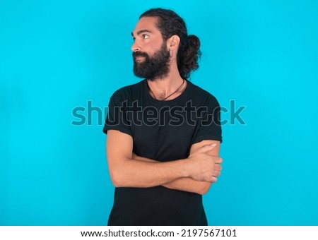 Pleased young bearded man wearing black T-shirt over blue studio background keeps hands crossed over chest looks happily aside
