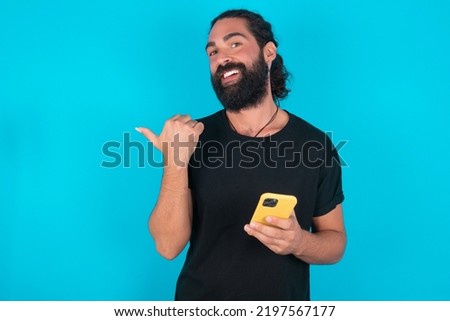 young bearded man wearing black T-shirt over blue studio background using and texting with smartphone  pointing and showing with thumb up to the side with happy face smiling