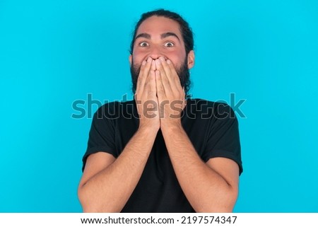 Vivacious young bearded man wearing black T-shirt over blue studio background, giggles joyfully, covers mouth, has natural laughter, hears positive story or funny anecdote Royalty-Free Stock Photo #2197574347