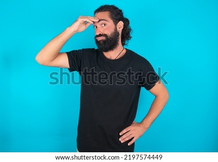 young bearded man wearing black T-shirt over blue studio background having problems, worried and stressed holds hand on forehead.
