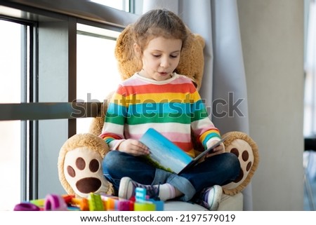 A cute girl of 6 years old sits on the couch by the window with a large toy bear and reads a book. She is dressed in a colored jumper. Kids. Teaching. Royalty-Free Stock Photo #2197579019