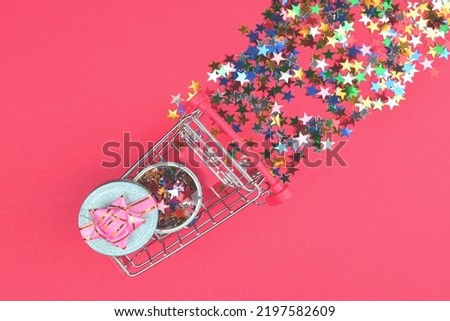 Blue present box with pink bow in a shopping cart and confetti on a red background. 