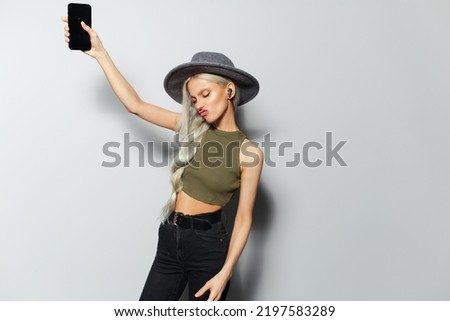 Studio portrait of beautiful young happiness blonde girl, dancing and listen the music with wireless earphones, holding smartphone in hand, wearing grey hat on white background.