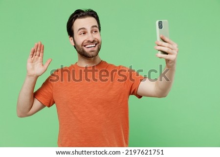 Young fun man 20s wear orange t-shirt doing selfie shot on mobile cell phone post photo on social network waving hand say hello isolated on plain pastel light green color background studio portrait.