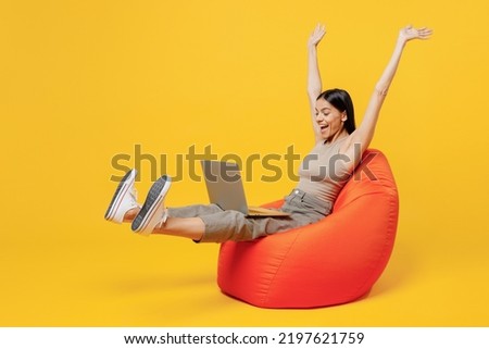 Full body young overjoyed happy latin woman 30s she wear basic beige tank shirt sit in bag chair hold use work on laptop pc computer spread raise up hands isolated on plain yellow backround studio.