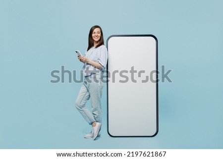 Full size young happy woman she 20s in casual blouse big huge blank screen mobile cell phone with workspace copy space mockup area hold smartphone isolated on pastel plain light blue background studio