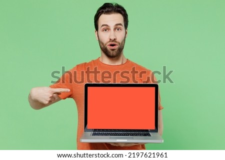 Young amazed fun man 20s wearing orange t-shirt hold use work point index finger on laptop pc computer with blank screen workspace area isolated on plain pastel light green color background studio.