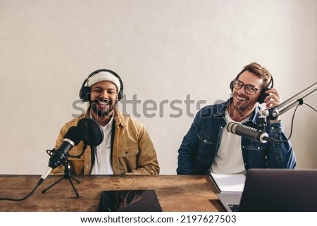 Two happy radio presenters having a good time on air. Young men smiling happily while recording an audio broadcast in a studio. Cheerful content creators co-hosting an internet podcast. Royalty-Free Stock Photo #2197627503