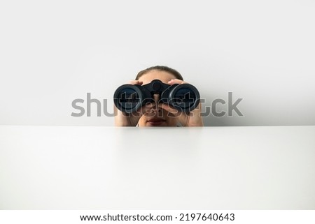 Young woman looking through binoculars peeking out from behind white cardboard on white background.
