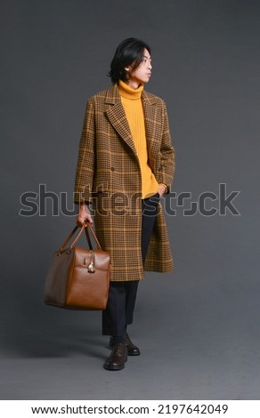 fashion model. full body Young man with hairstyle in striped, coat with yellow sweater holding handbag  posing on gray background Royalty-Free Stock Photo #2197642049