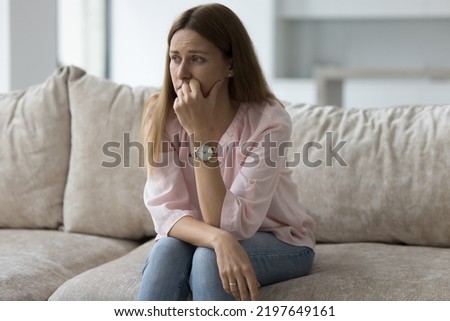 Unhappy depressed pretty woman sitting on sofa at home, looking away, thinking over health problems, crisis, loss, bad news, touching face, feeling stressed, frustrated. Negative emotions Royalty-Free Stock Photo #2197649161