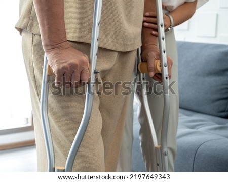 Closeup elderly woman's hands holding on crutches standing in living room at home, trying to walk. Helping and care senior patient to walk with crutches walker concept.