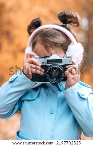 Cute little caucasian girl in a blue raincoat and fur headphones takes pictures with a camera in an autumn park.Autumn concept.
