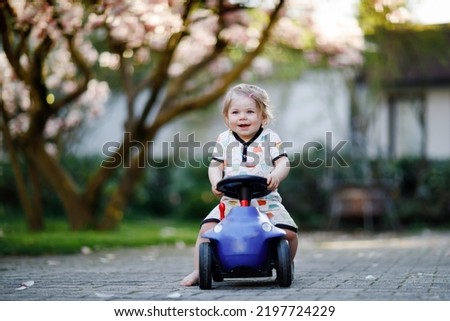Cute little baby girl playing with blue small toy car in garden of home or nursery. Adorable beautiful toddler child with blossoming magnolia on background