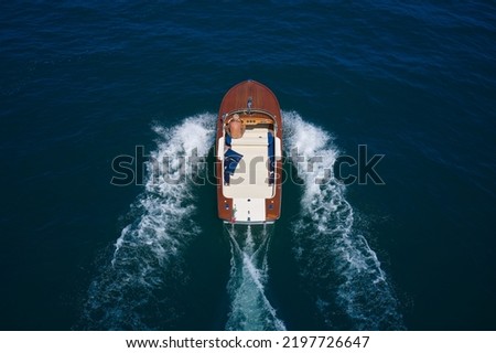 Wooden expensive boat fast movement on dark water top view. Big expensive wooden open boat with a man moving fast on dark blue water top view. Italian wooden speedboat moving up aerial view. Royalty-Free Stock Photo #2197726647
