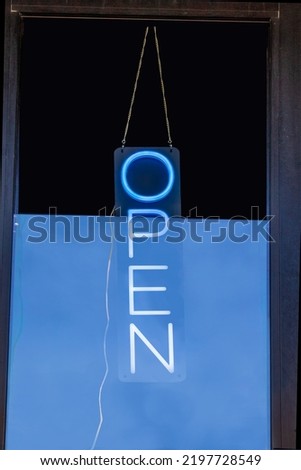 Neon OPEN sign hanging on door to business with blue sky reflected in glass door and electric cord dangling from sign