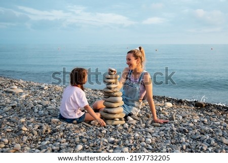 A mother and daughter 6 years old on the beach build a castle of stones. They socialize and have fun together. Family. Summer. Vacation.