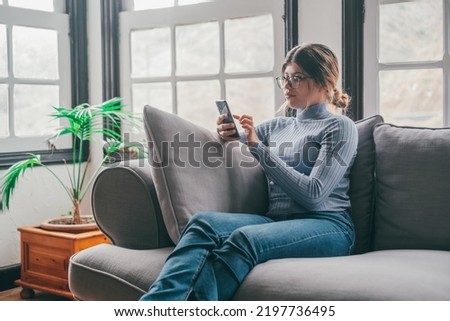 Young beautiful woman sitting on the sofa at home chatting and surfing the net. Female person having fun with smartphone online. Portrait of girl smiling using cellphone revising social media