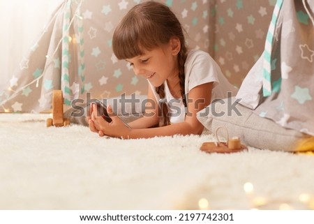 Side view portrait of smiling little girl with braids and dark hair wearing white t shirt posing in wigwam, child using mobile phone with interest, expressing positive emotions.