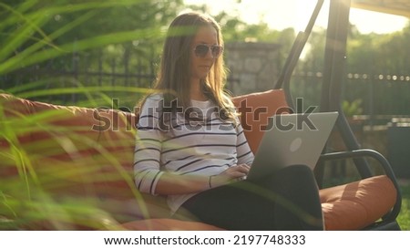 Woman sitting in swinging couch outdoor in garden working with laptop computer. Home office, working on vacation, sun light, lens flare. Royalty-Free Stock Photo #2197748333