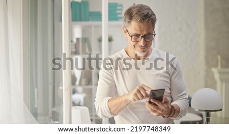 Happy older good looking casual man using phone at home, smiling. Portrait of mid adult, mature age man in glasses, Royalty-Free Stock Photo #2197748349