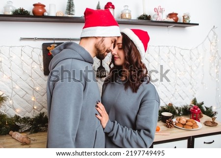 Lovely young romantic couple standing kitchen wear red hats kissing garlands home cosy interior atmosphere New Year Christmas tree decorations holiday party celebrating concept winter evening 
