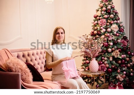 Charming woman dressed in a white wool suit holds a pink gift, against the background of a decorated Christmas tree in a room next to a New Year tree and a pink sofa, gifts and candles