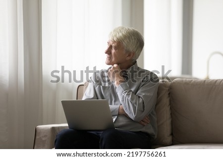 Thoughtful dreamy mature elder man keeping laptop on lap, looking at window away, thinking, daydreaming, touching chin, relaxing at home. Internet, online communication, retirement