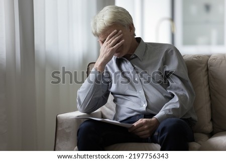 Desperate depressed senior elder man getting bad shocking news from paper documents, sitting on sofa at home, covering face with hand, receiving concerning medical result, bankruptcy notice Royalty-Free Stock Photo #2197756343
