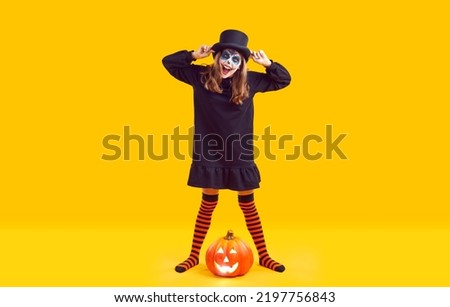 Preteen girl with creative make up and in Halloween costume having fun on orange background. Full length child in black dress, hat and orange and black leggings standing near pumpkin and laughing.
