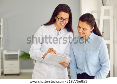 Friendly nurse holding clipboard and communicating with patient. Happy doctor interviewing young woman, filling out questionnaire with personal details, or showing her history record and exam results Royalty-Free Stock Photo #2197756853