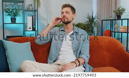 Happy relaxed overjoyed young guy in wireless earphones dancing on couch at home choosing listening favorite energetic disco rock n roll music in smartphone entertaining. People weekend activities