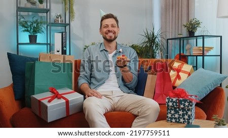 Happy adult man wears festive birthday hat hold cupcake makes wish joyful congratulating blowing burning candle on cake. Young guy celebrating anniversary party at home room apartment on orange couch Royalty-Free Stock Photo #2197759511