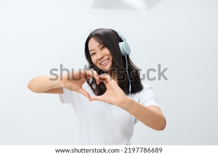Happy smiling cute Asian young female in white basic t-shirt show heart shaped gesture at camera posing isolated on over white studio background. The best offer for ad. Favorite Music App Ad concept