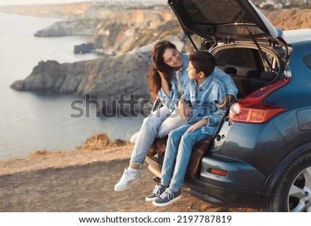 Family trip to the mountains by car. Mom is having fun with her teenage son, sitting in the trunk of a car and enjoying the view together.