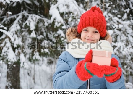 cute teen girl opens a gift box, christmas present, outdoor in winter, child in scarf, mittens and hat, snow and winter park on background