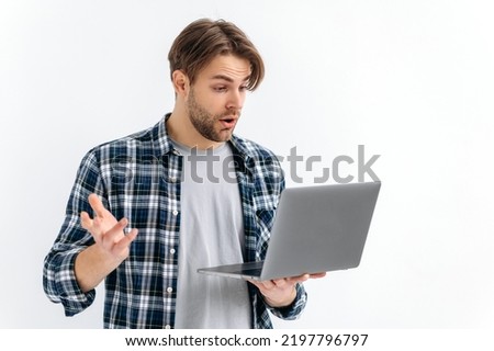 Shocked confused puzzled caucasian stylish young man, standing on a white isolated background, holds an open laptop, surprised looks at the screen, see unexpected news, message, gesturing with hand