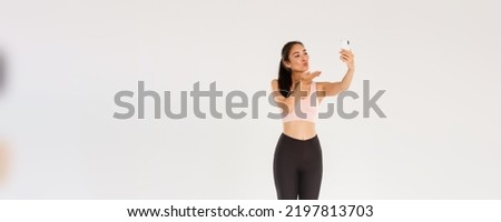 Full length of cute and silly female blogger in gym, wearing sportswear and taking selfie or live stream from gym workout session, sending air kiss at mobile phone screen, standing white background
