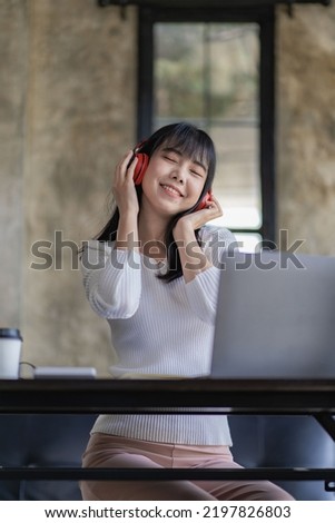 Asian woman sitting at a table and connected to a laptop She wears headphones and enjoys listening to music at work.