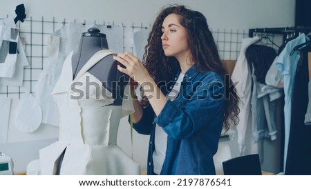 Young female tailor is adjusting clothes on tailoring dummy with sewing pins and measuring with measure-tape. Women's garments, sketches on wall, tailoring items and tools are visible. Royalty-Free Stock Photo #2197876541