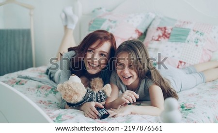 Laughing happy mother and cute daughter watching funny cartoon movie on TV while lying on bed at home in morning have fun at cozy bedroom