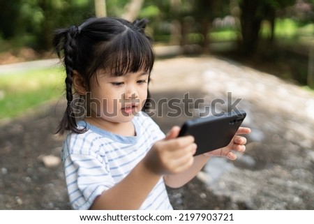 Asian baby girl smiling and using mobile selfie make a photo picture in the park garden. Cute girl learning to use smartphone by her self. Education learning baby concept.
