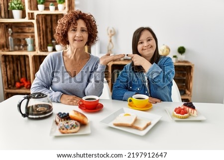 Family of mother and down syndrome daughter sitting at home eating breakfast amazed and smiling to the camera while presenting with hand and pointing with finger. 