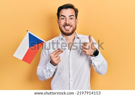 Handsome caucasian man with beard holding czech republic flag smiling happy and positive, thumb up doing excellent and approval sign 
