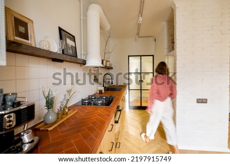 Stylish kitchen interior of modern apartment with motion blurred female person walking inside. Interior made in white and beige tones with tiled table top and glass door