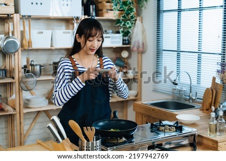 portrait of a cheerful Chinese girl using her smartphone to take pictures of the food in the frying pan while cooking in a rustic kitchen with daylight at home.